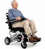 A product image of the Air Hawk Foldable Power Wheelchair – Portable, ideal for travel. An older man smiling while seated in a compact electric wheelchair with a joystick, photographed on a clean white studio background. The wheelchair frame is silver with black wheels and seat, and the brand name AIR HAWK is visible on the side.